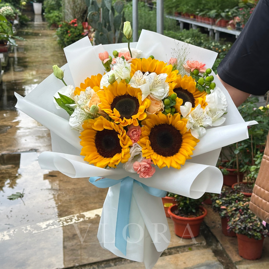 Bouquet of sunflowers and other flowers with a blue ribbon, held by a person in an outdoor setting.