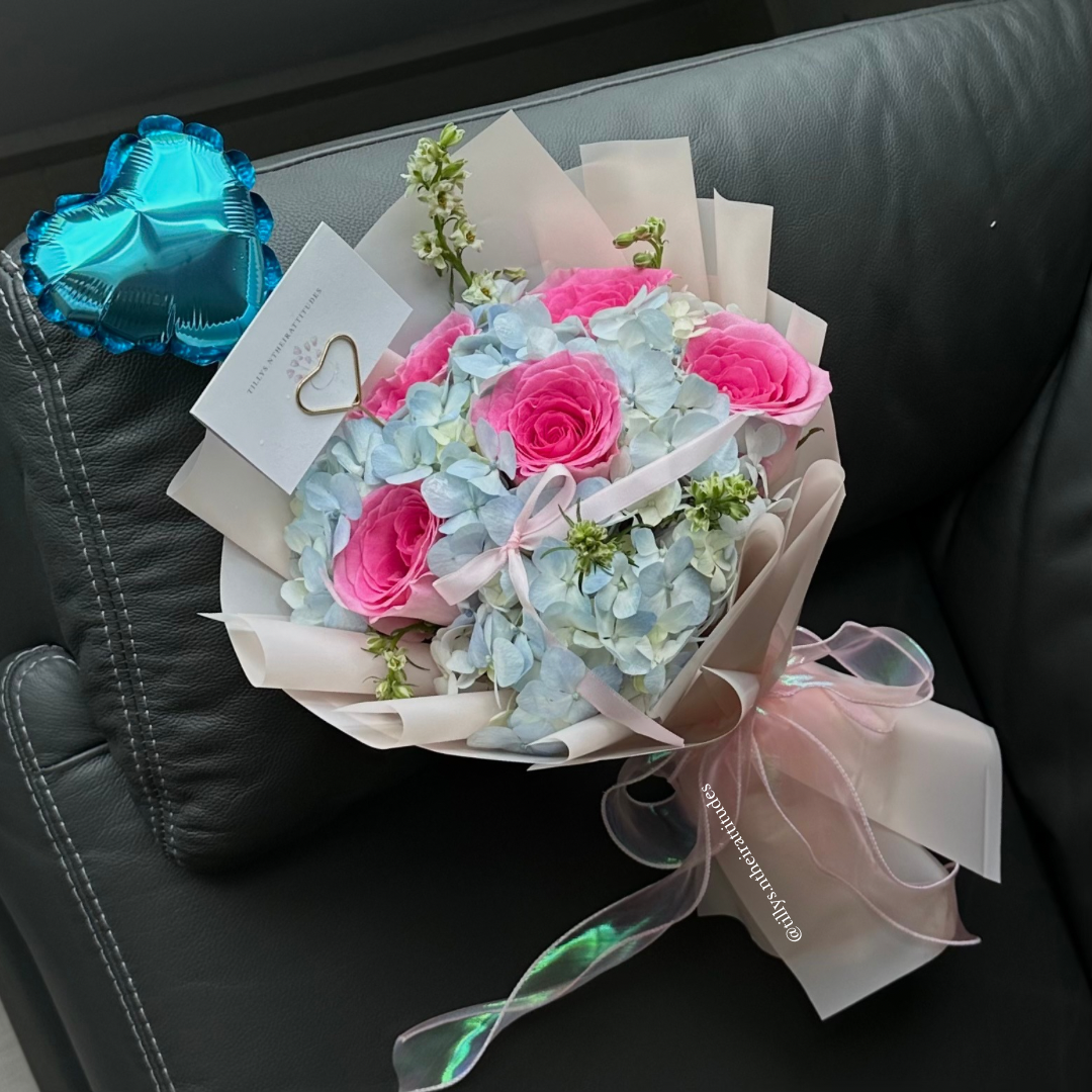 A hand bouquet with pink roses and blue hydrangea, tied with light pink ribbons and a shimmering blue chrome balloon in the background.