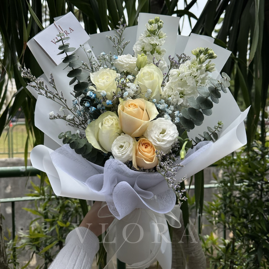 Bouquet of white and yellow roses with greenery, held by a person wearing gloves, with a blurred background.