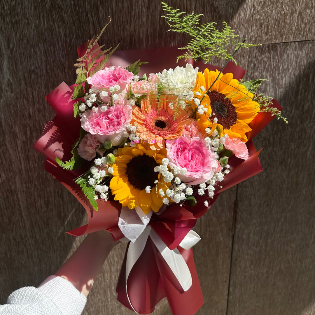 Bouquet of flowers with pink roses, yellow sunflowers, and greenery against a wooden background