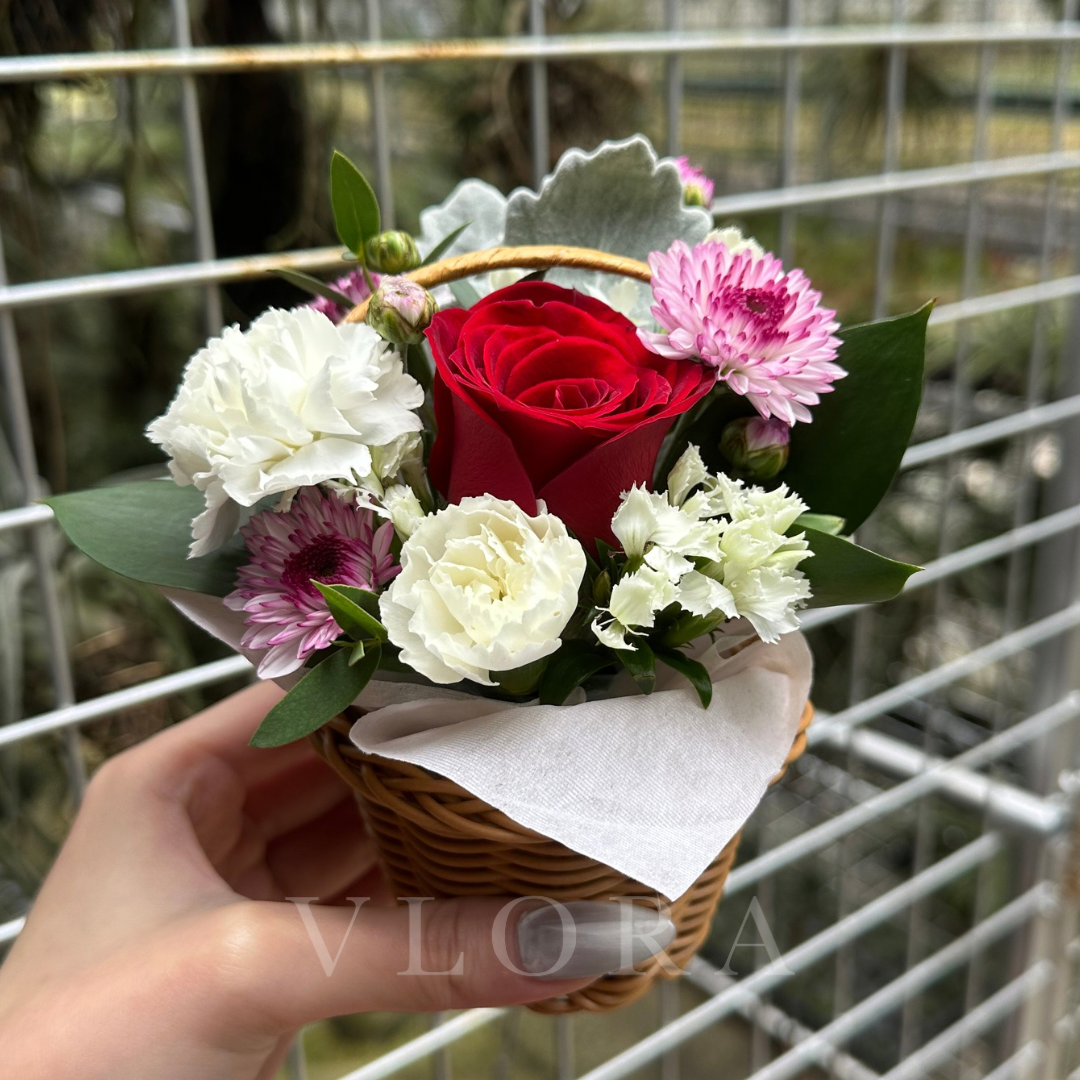 Small bouquet of flowers in a basket held by a hand with a blurred metal fence background