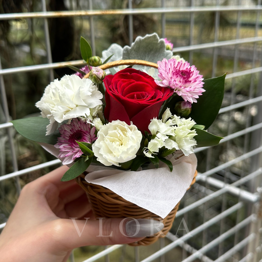 Small bouquet of flowers in a basket held by a hand with a blurred metal fence background