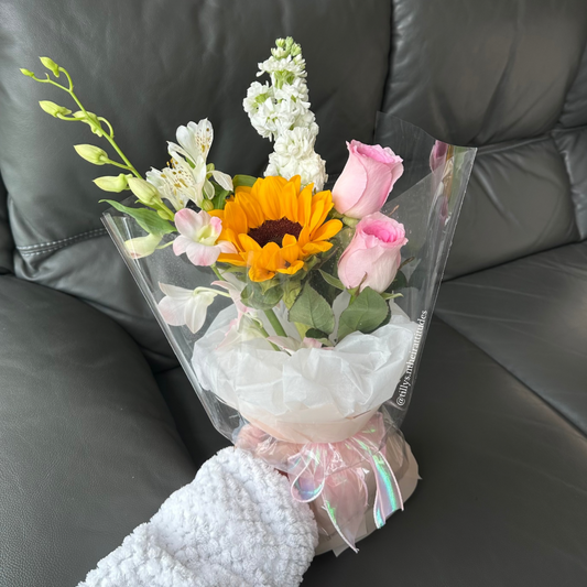 A petite bouquet consisting of a bright Sunflower, two soft pink Roses, a light pink Orchid, and gentle side blooms, wrapped in a clear plastic with a decorative ribbon.