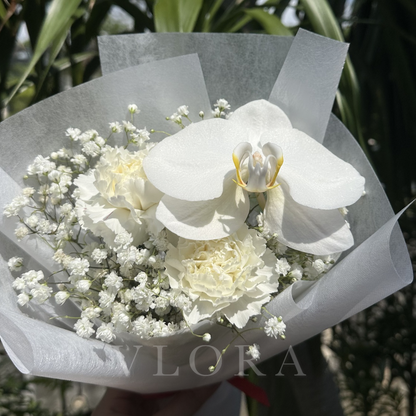 A bouquet of white flowers including a large white orchid, white carnations, and baby's breath, wrapped in translucent white tissue paper and held outdoors with a hand visible at the bottom.