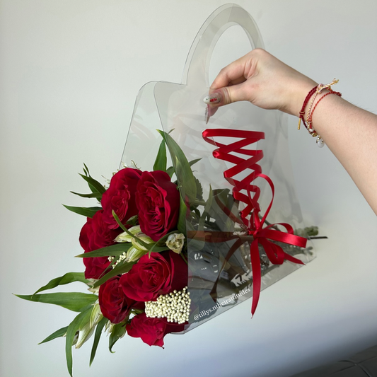 A floral tote bag arrangement featuring red flowers and green leaves, tied with a red ribbon, being held by a person.