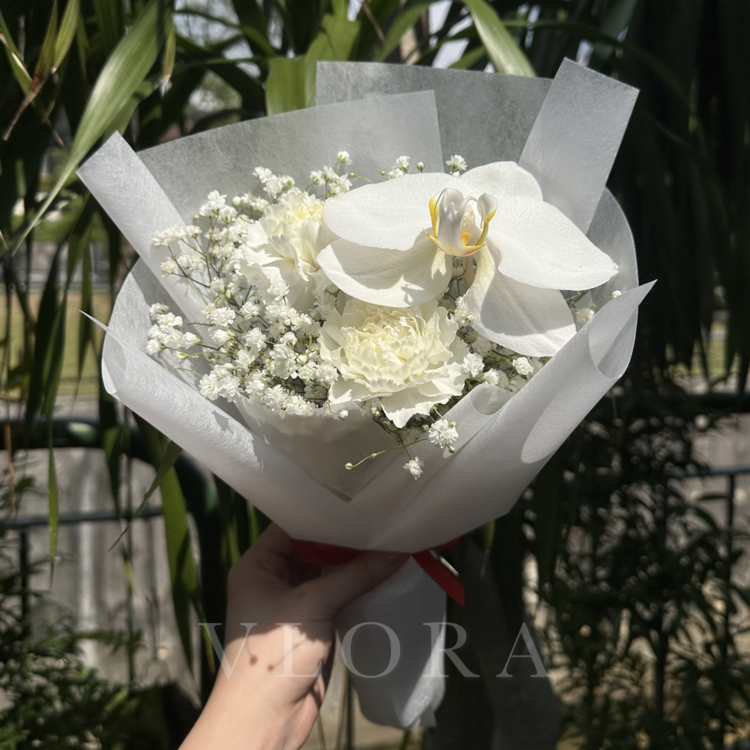 A hand holding a bouquet of white flowers, including a large white orchid, cream carnation, and baby's breath, wrapped in white tissue paper with a red ribbon, set against a blurred background of green foliage.