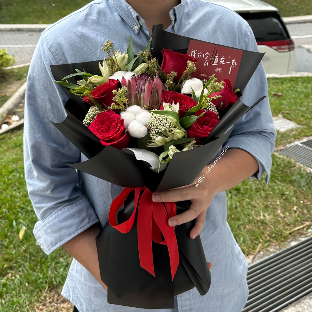A person holding a bouquet of red roses, white flowers, and black wrapping with a red ribbon.