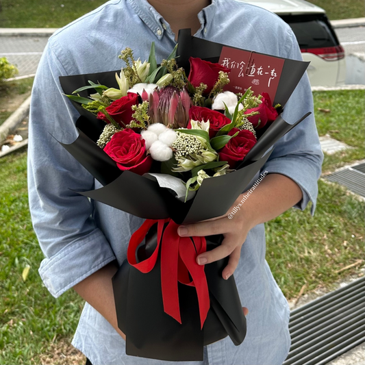 A person holding a bouquet of red roses, white flowers, and black wrapping with a red ribbon.