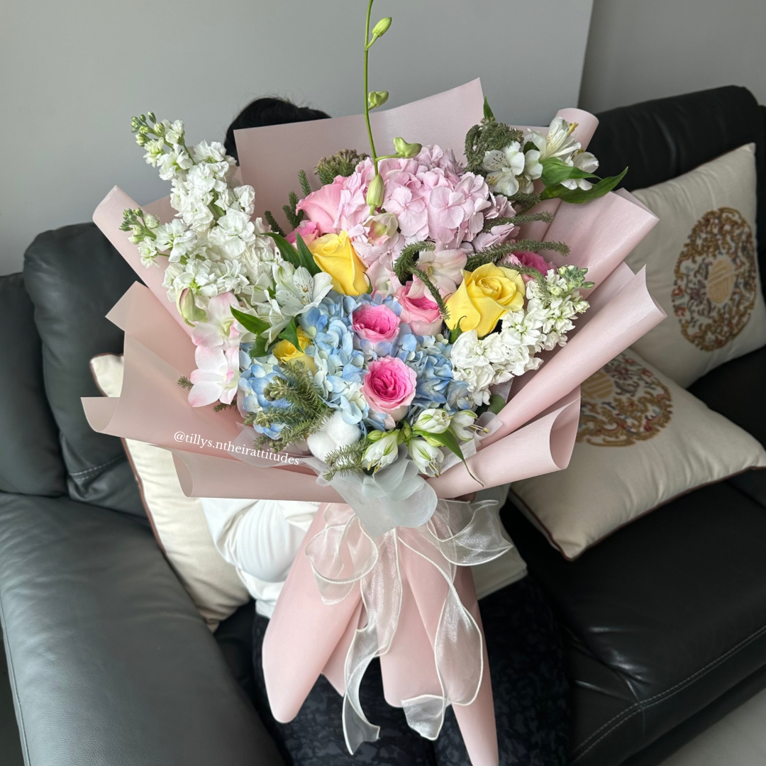 A bouquet of flowers featuring blue & pink hydrangeas, yellow Roses, and white Mattiolas, wrapped in white paper with an organza white ribbon, placed on a sofa.