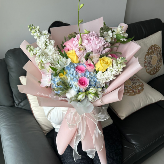 A bouquet of flowers featuring blue & pink hydrangeas, yellow Roses, and white Mattiolas, wrapped in white paper with an organza white ribbon, placed on a sofa.