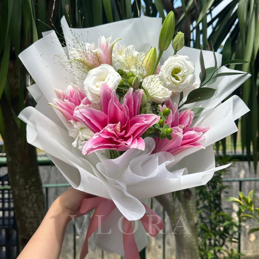 Bouquet of pink and white flowers with a white wrap held by a person, with a blurred green background.