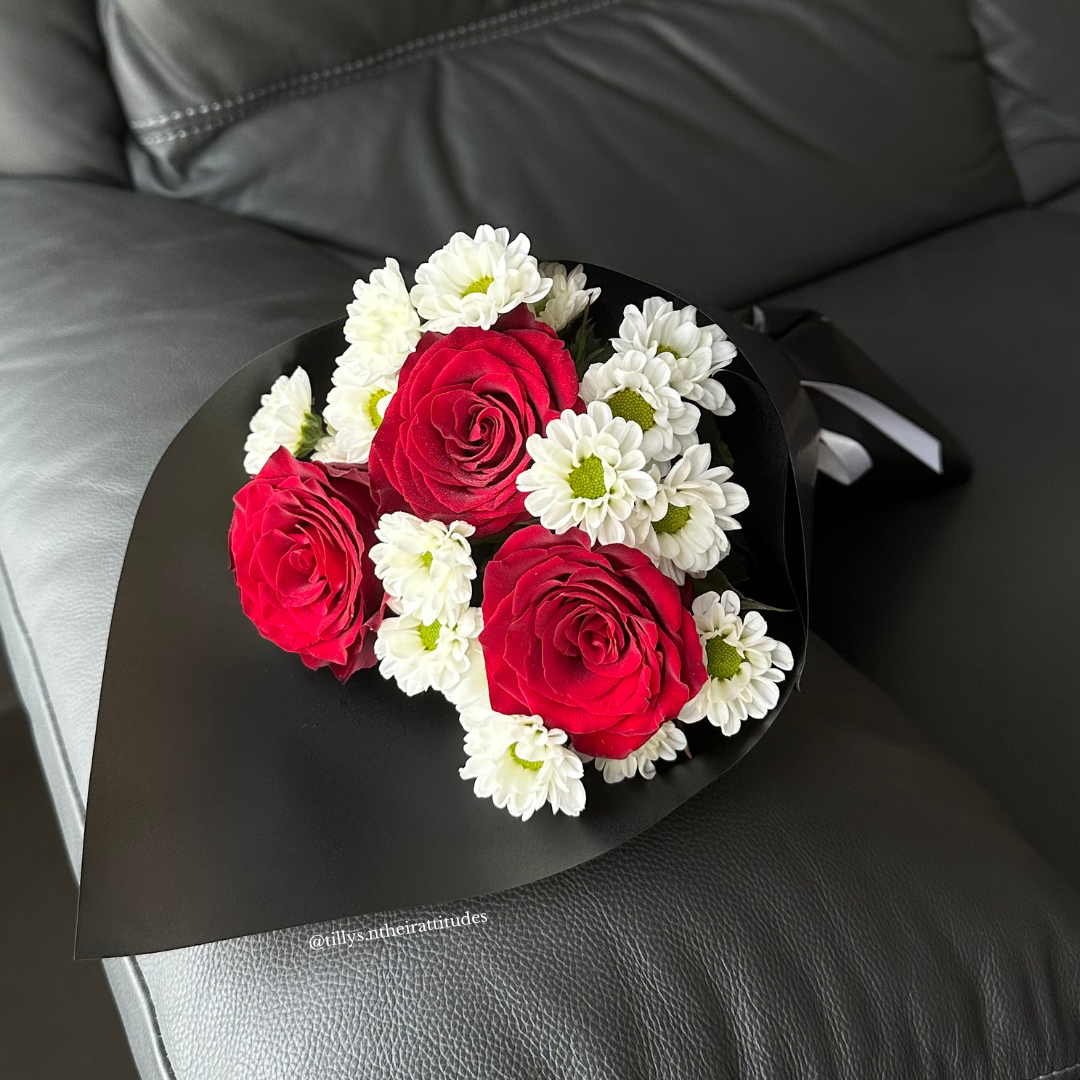 A bouquet of red roses and white Chrysanthemums wrapped in black paper, placed on a leather surface.