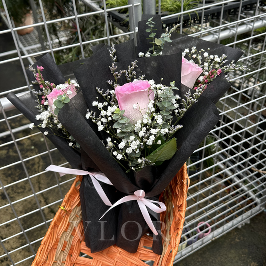 Bouquet of pink roses and greenery wrapped in black paper with a pink ribbon, placed on an orange woven basket.