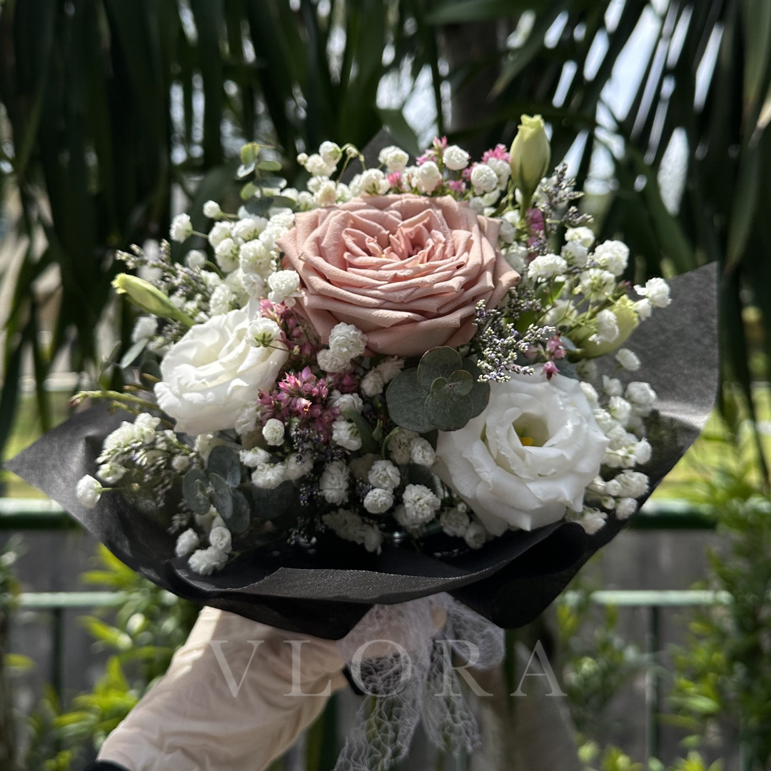 Bouquet of mocha Rose flowers (table arrangement) held by a person with 'Vlora' branding, against a blurred green background.