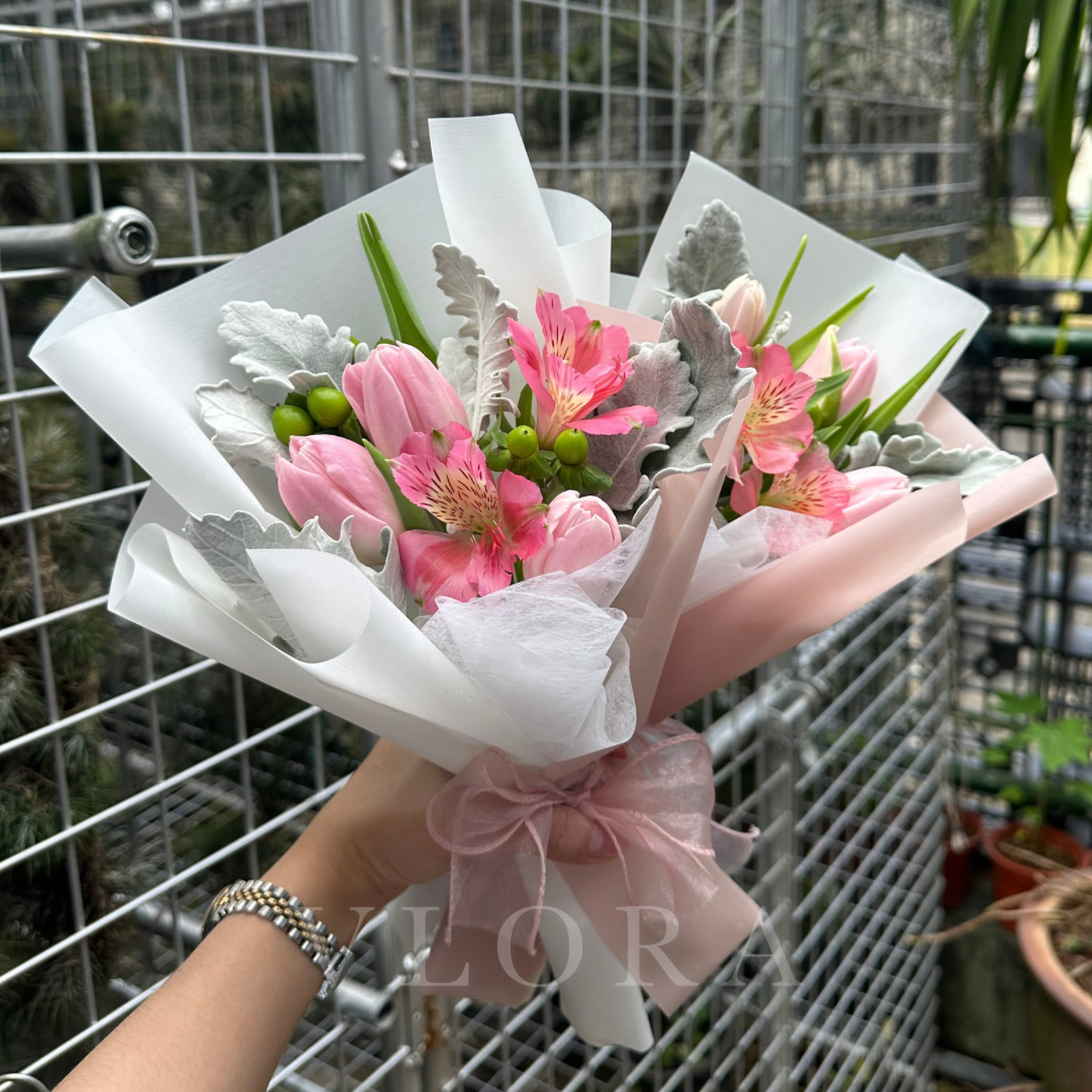 Bouquet of pink tulips and mixed flowers with greenery wrapped in white paper and a pink bow, held by a person.