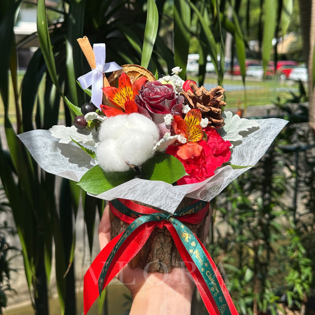 A table arrangement of Christmas blooms in a wooden bark pot with a red rose, white cotton flower, pine cones, lemon slices and a cinnamon stick.