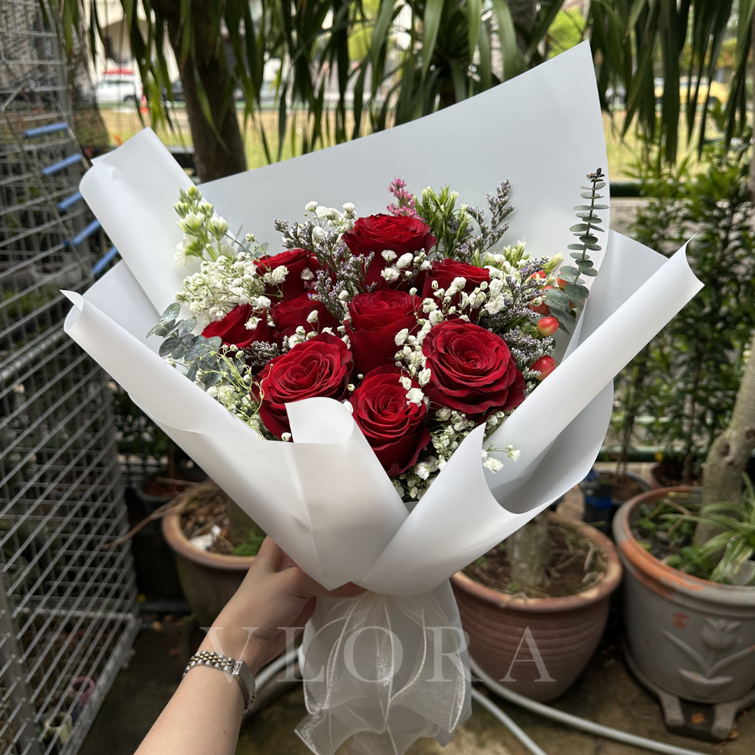 Bouquet of 9 red roses with greenery wrapped in white paper, held by a person in an outdoor setting.